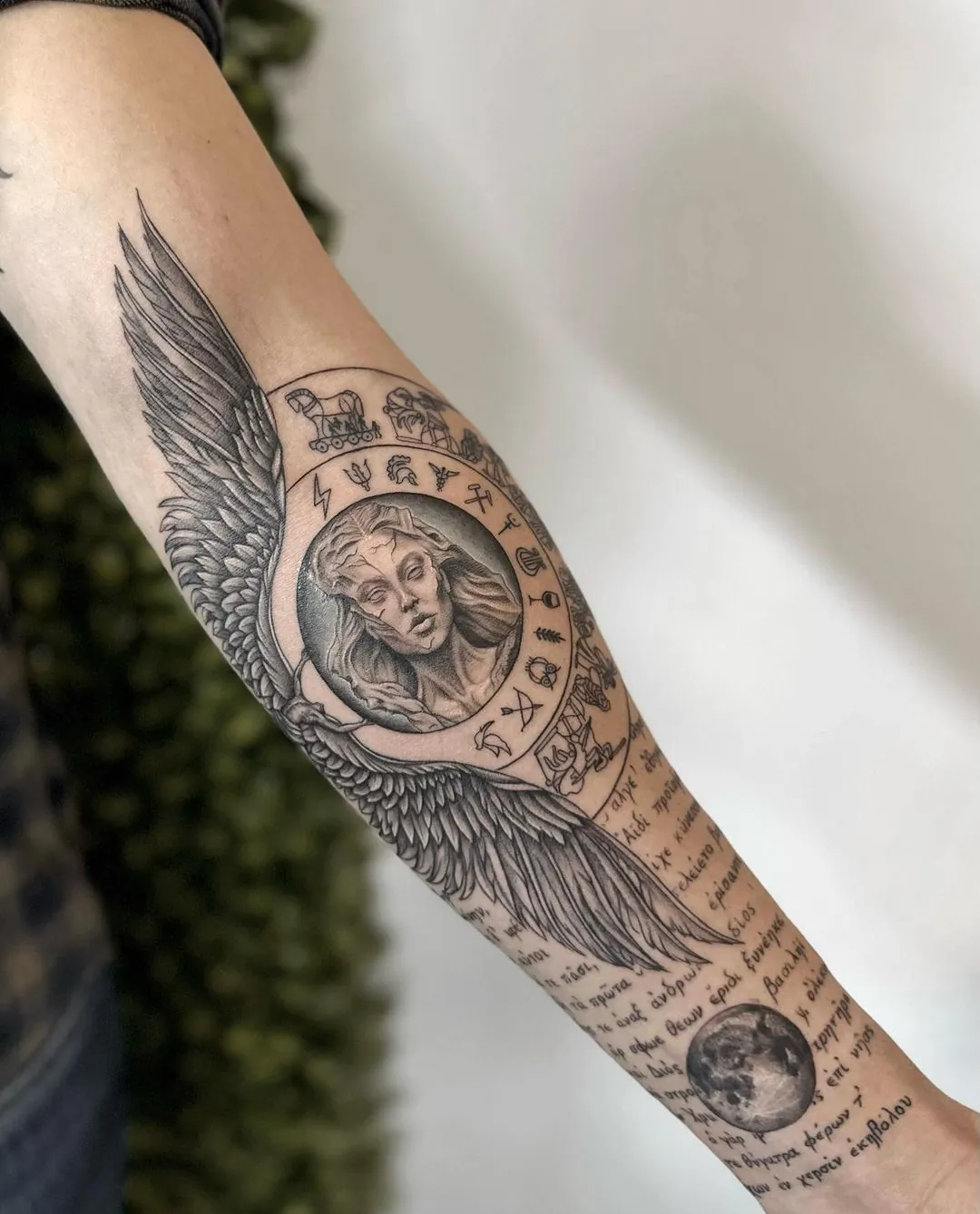 Tatuaj alb-negru pe antebraț cu aripi, medalion și text