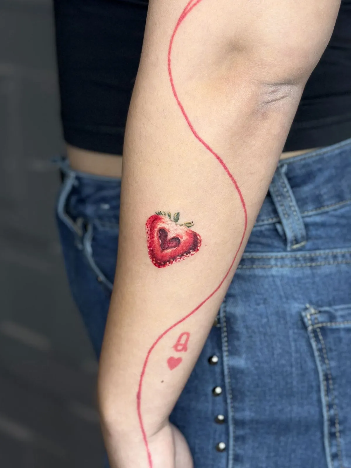 Tatuaj color pe braț cu o căpșună tăiată cu o inimă în interior, unită printr-un fir roșu de un simbol de carte de joc