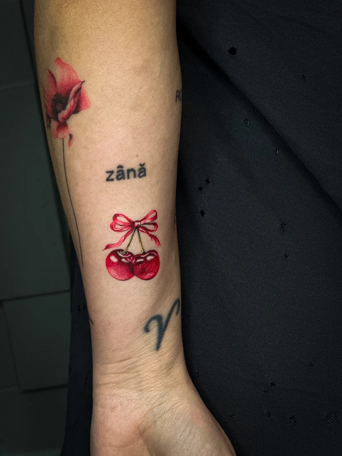 Tatuaj color pe antebraț cu cireșe legate cu o fundă roșie și textul zână