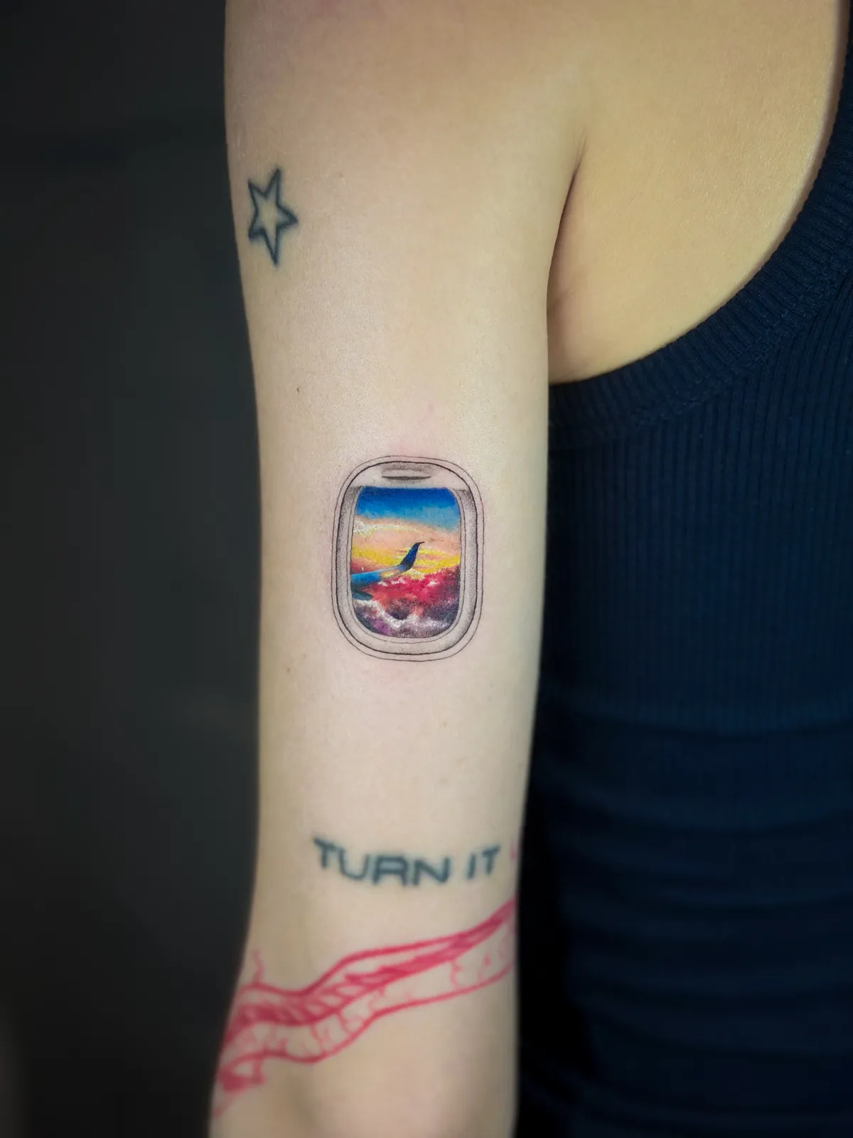 Tatuaj color pe braț cu un hublou de avion și un apus înflăcărat printre nori