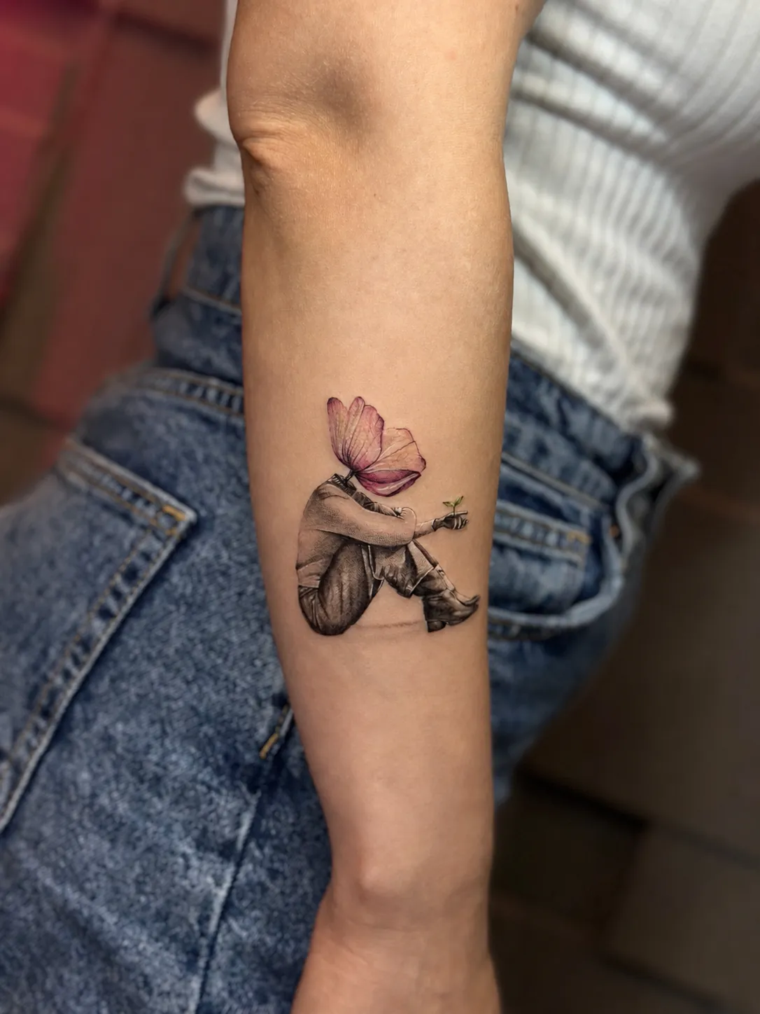 Tatuaj fine line pe antebraț cu o siluetă fără cap, cu cap de fluture roz, ținând un mugur verde