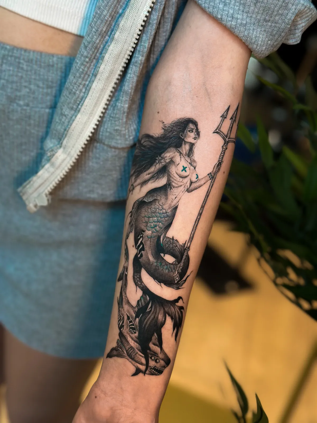 Tatuaj alb-negru pe antebraț cu o sirenă cu trident, luptând cu un rechin