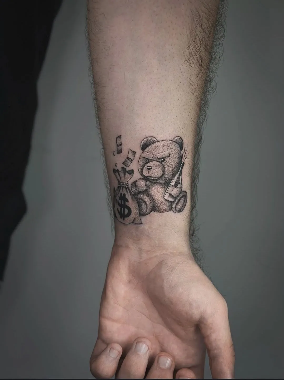 Tatuaj alb-negru pe antebraț cu un ursuleț, un sac de bani și o sticlă de șampanie