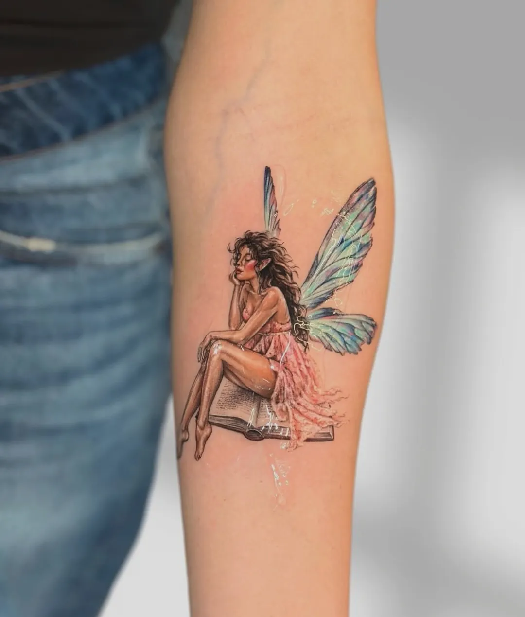 Tatuaj color pe antebraț cu o zână așezată pe o carte deschisă